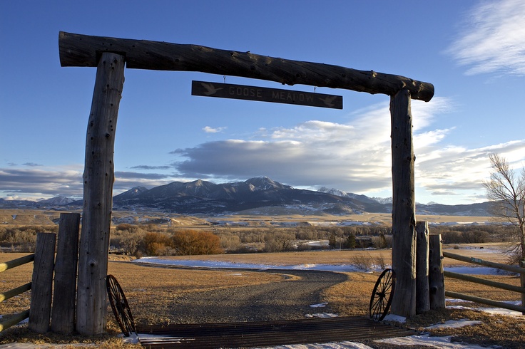 A log arch with a gravel road through it before a field and Livingston Peak on a clear, autumn day.