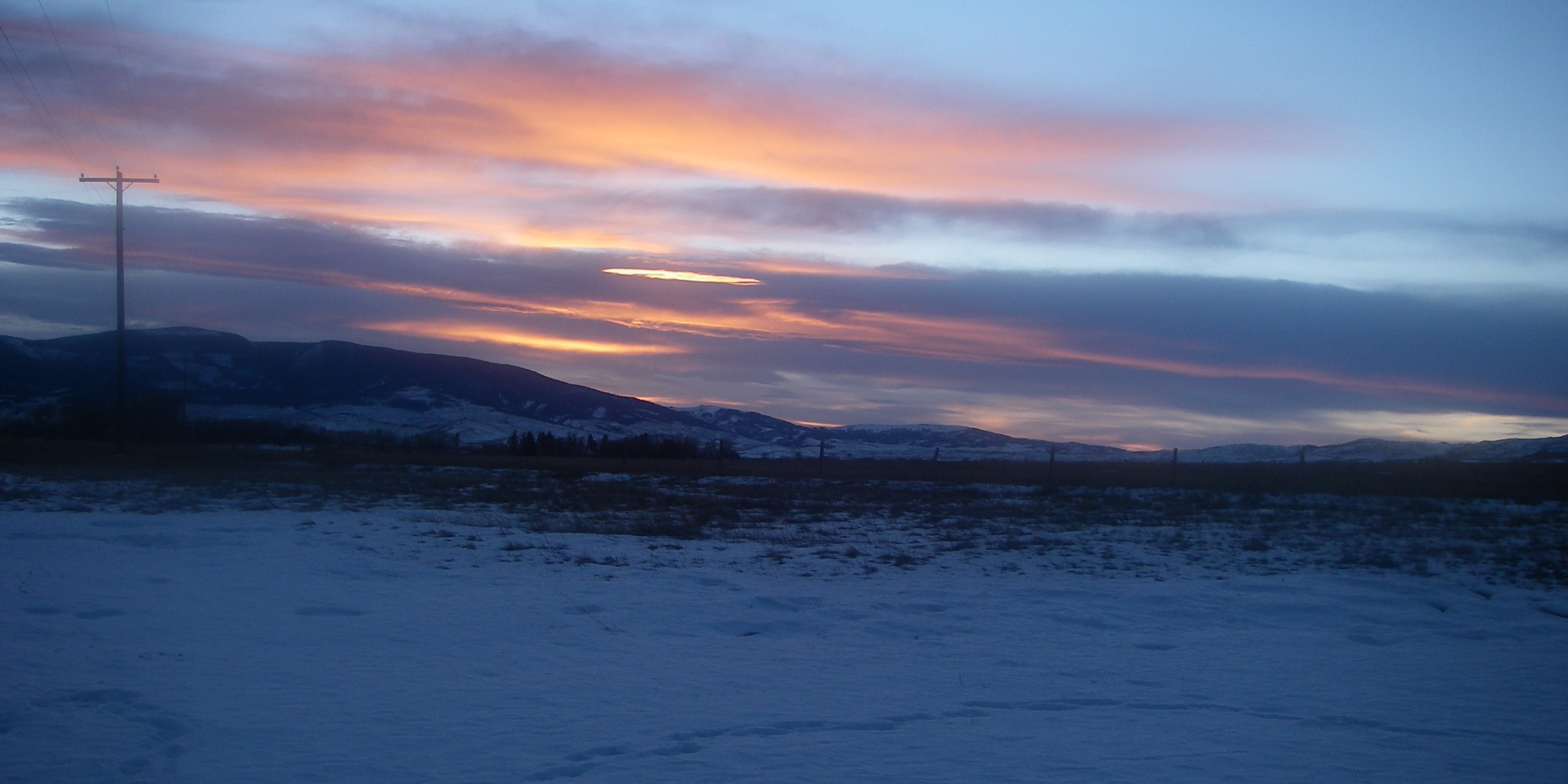 A snowy field and a telephone pole on the left at sunset, with orange clouds above the outline of the mountains